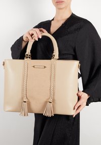 Beige håndtaske med vævet tekstur, fletteaccenter og frynser. Har et struktureret design og dobbelte top håndtag. Detaljer i guldfarve.