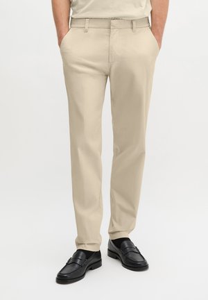 Beżowe spodnie o prostym kroju noszone z czarnymi skórzanymi loafersami i czarnymi skarpetkami, ręce w kieszeniach na jednolitym jasnym tle.