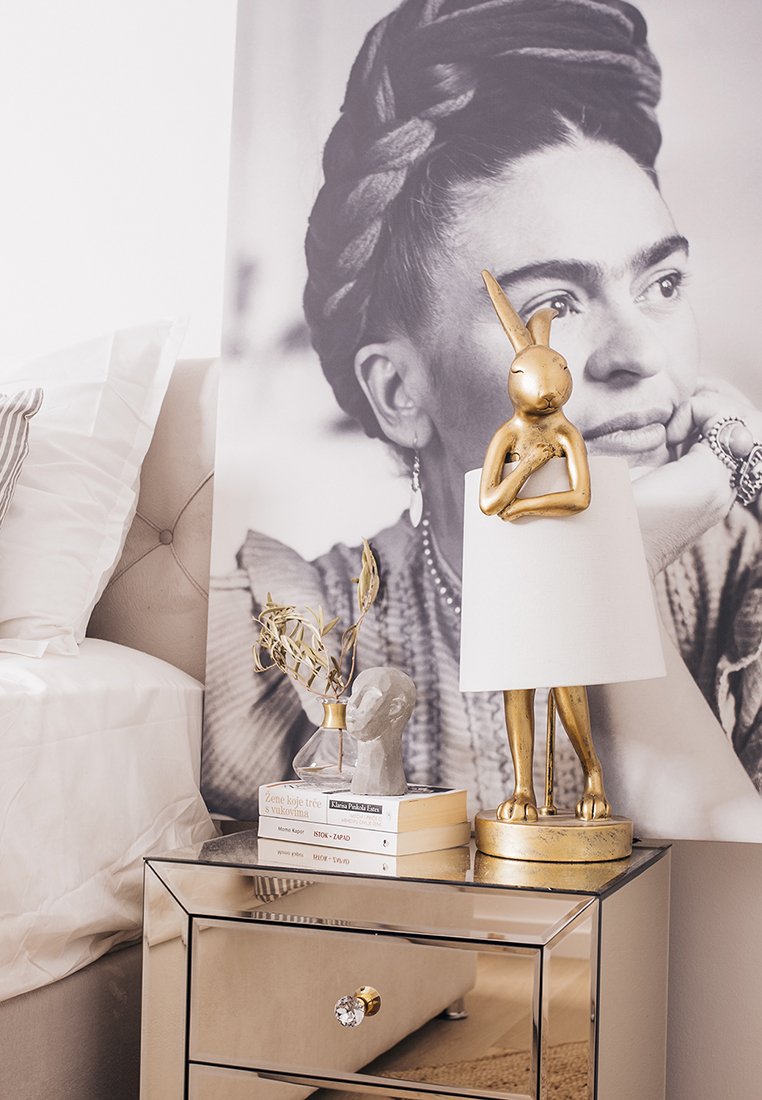 Goldfarbener Hase-Lampe mit weißem Schirm, grauem Dekorationsobjekt und gestapelten Büchern auf einem verspiegelten Nachttisch neben einem schwarz-weißen Porträt.