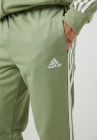 Maslinasto zelene Adidas trenirke hlače s bijelim trostrukim prugama, glatke teksture, uskog kroja i s malim izvezenim logotipom na bedru.