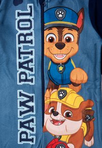 Tuta in pile a tema Paw Patrol, realizzata in materiale blu, con personaggi animati e una cerniera. "PAW PATROL" in evidenza in lettere bianche.