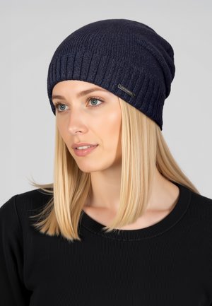 Blonde woman wearing a dark blue knit beanie and black sweater, facing slightly right against a plain light gray background.