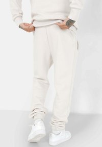 Lichtbeige joggingsbroek met elastische boorden en zijzakken. Zachte stoftextuur, ontspannen pasvorm. Draagt bij witte sneakers en met een zichtbare geribbelde tailleband.