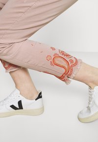 Pantalones cortos de color rosa claro con bordados de paisley rojos y dobladillos con flecos, combinados con zapatillas blancas que tienen detalles en negro y suelas texturizadas.