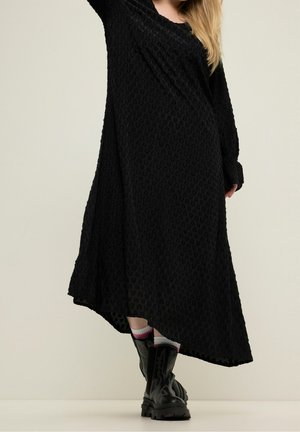 Frau trägt ein langes schwarzes strukturiertes Kleid und schwarze klobige Stiefel mit weißen und pinkfarbenen Socken, steht vor einem einfarbigen hellen Hintergrund.