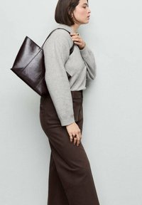 Mujer que lleva un suéter gris y pantalones marrones, con un bolso tote de cuero marrón oscuro sobre el hombro, mirando hacia un lado.