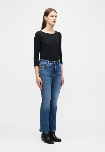 Femme debout, portant un haut noir à manches trois-quarts, un jean bleu droit, et des bottines noires sur fond blanc.