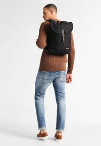 Svart tygryggsäck med rullstängning, en rem och spänne detaljer, samt en logotyp. Bärs av en person med en brun tröja och blå jeans.