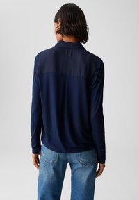Dunkelblaues Langarmhemd mit einem glatten, satinartigen Oberteil und einem strukturierten unteren Abschnitt; kombiniert mit hellblauen Jeans.