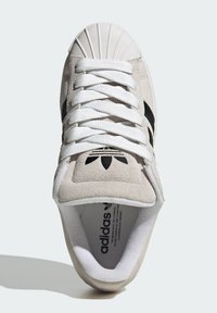 Béžová semišová teniska s bielymi šnúrkami, dizajnom s chráničom na špičke a tromi čiernymi pruhmi. Vyznačuje sa textúrovaným vrchom a detailom loga.