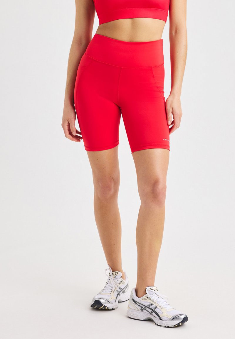 Mujer con pantalones cortos de ciclismo de talle alto rojos brillantes y sujetador deportivo, de pie con zapatillas de running blancas sobre un fondo neutro.