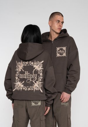 Hættezip-sweater i mørkebrunt stof med en beige grafisk design på ryggen og fronten. Akkompagneret af cargobukser i samme farve.