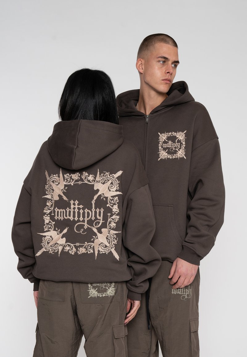 Hættezip-sweater i mørkebrunt stof med en beige grafisk design på ryggen og fronten. Akkompagneret af cargobukser i samme farve.