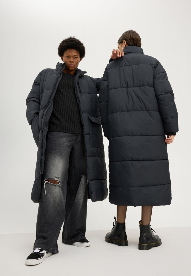 Abrigo acolchado oscuro con un patrón acolchado, que presenta un cuello alto y bolsillos laterales, combinado con jeans holgados y botas negras.