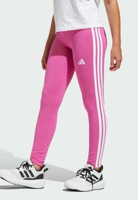 Leggings rosa con tre strisce bianche sui lati, caratterizzati da un logo Adidas sulla coscia sinistra. Il materiale sembra liscio e elastico.