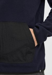 Marineblauw fleece pullover met een zwarte gestructureerde zak. Beschikt over een relaxte pasvorm en een contrasterende zoom. Zacht materiaal met een gladde afwerking.