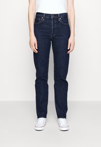 Person wearing dark blue straight-leg jeans, white sneakers, and a white top, standing against a plain light background.