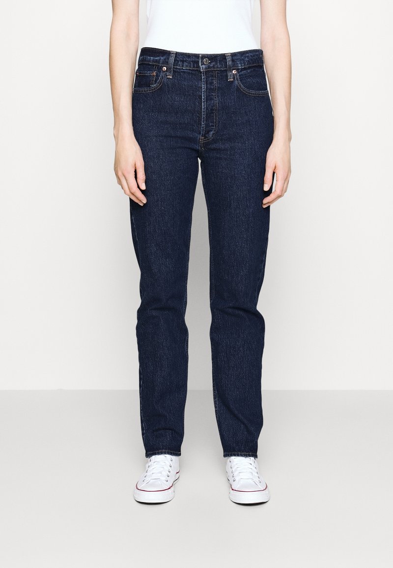 Person wearing dark blue straight-leg jeans, white sneakers, and a white top, standing against a plain light background.