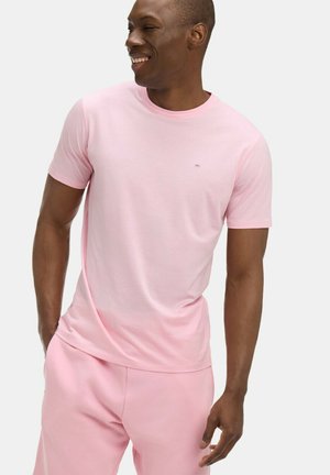Homme souriant portant un t-shirt à manches courtes rose clair et un pantalon rose assorti, debout avec une main dans la poche sur un fond blanc.