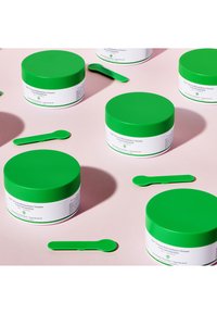 Grüne runde Gläser mit weißen Etiketten und grünen Akzenten, die einen Makeup-Reinigungsbutter enthalten, begleitet von passenden grünen Löffeln auf einem rosa Hintergrund.