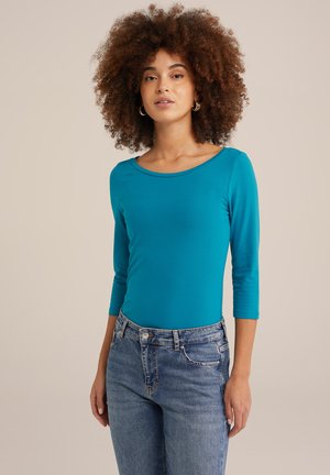 Turquoise lange mouwen top met een ronde halslijn, gedragen met lichtblauwe jeans. De stof is glad met een getailleerd ontwerp.