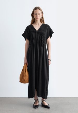 Femme debout, portant une longue robe noire plissée à col en V, des ballerines noires lacées, des boucles d'oreilles créoles et tenant un sac à main tressé marron.