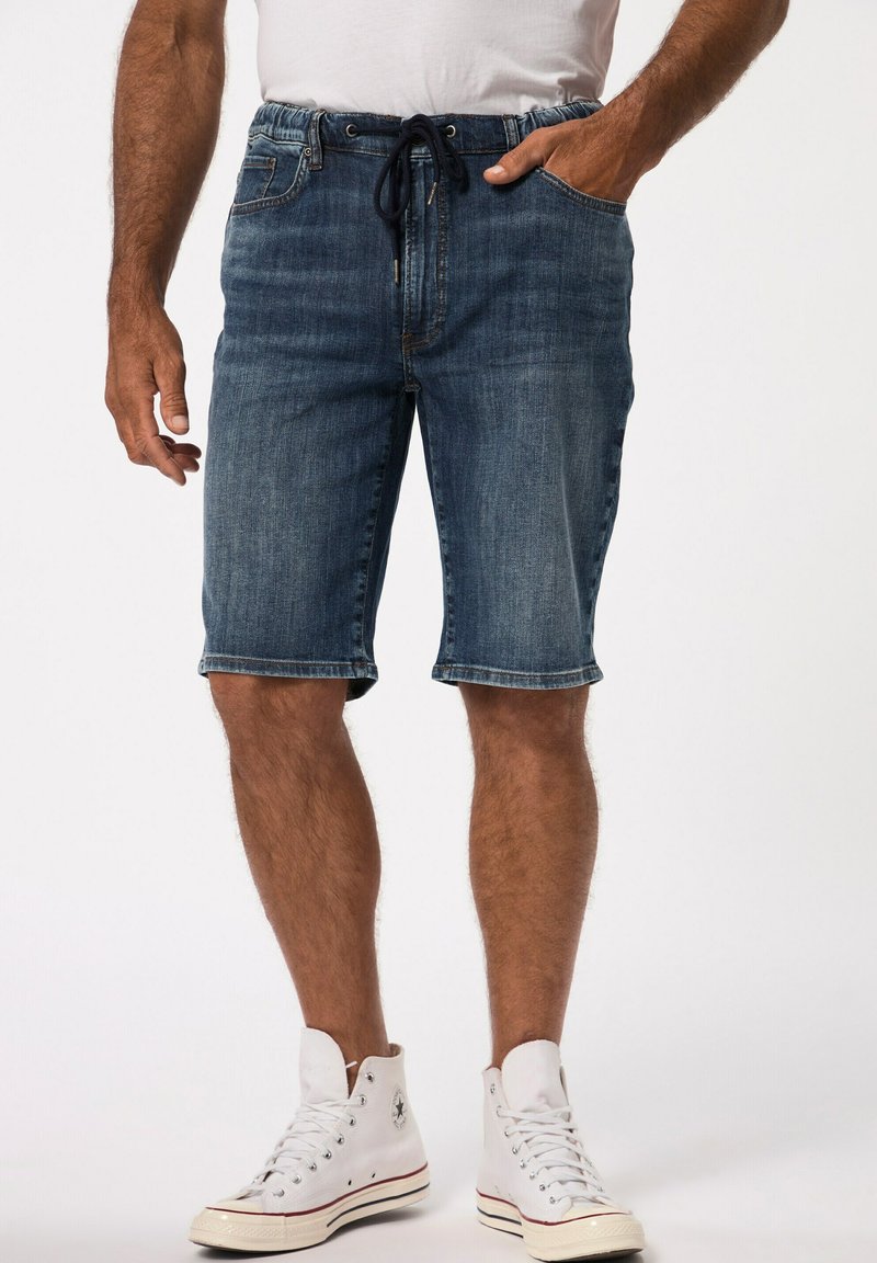 Shorts de mezclilla en azul oscuro con cinturilla ajustable. Presentan dobladillos enrollados, bolsillos delanteros y un ajuste relajado. Se llevan con zapatillas altas blancas.