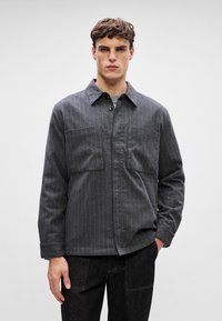 Szürke csíkos gombos zakó, két elülső zsebbel és gallérral, puha anyagból készült. Egy szürke, kerek nyakú póló fölött viselik.