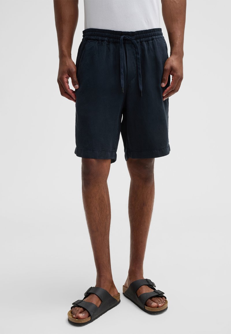 Pantaloni corti blu navy con vita elastica e cordoncino, caratterizzati da una vestibilità rilassata e orli arrotolati. Abbinati a sandali neri a due cinghie.