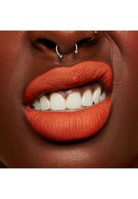 Matte orange läppfärg på fylliga läppar, som visar textur och detaljer. Vita tänder synliga, med en silverfärgad näspiercing. Mörk hudton.