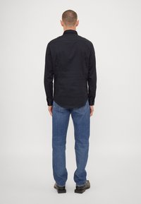 Camisa negra de manga larga con cuello clásico, corte recto y botones en los puños, combinada con jeans azules de pierna recta.