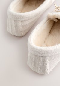 Chaussons doux couleur crème avec un motif torsadé tricoté et une doublure en peluche, vus de dessus sur une surface claire.