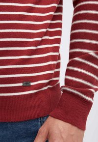 Pullover a righe rosse e bianche realizzato in morbido tessuto a maglia. Presenta un colletto e polsini a coste, con un piccolo tag metallico del logo sull'orlo.