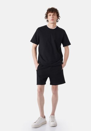 Jonge man staat met handen in zijn zakken, draagt een zwart T-shirt, zwarte korte broek en witte sneakers tegen een effen lichte achtergrond.