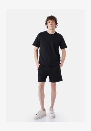 Jonge man staat met handen in zijn zakken, draagt een zwart T-shirt, zwarte korte broek en witte sneakers tegen een effen lichte achtergrond.