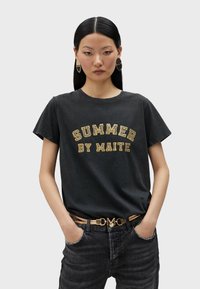 Camiseta negra con texto amarillo que dice "VERANO DE MAITE." Combinada con jeans oscuros y un cinturón dorado con un acento en forma de corazón.