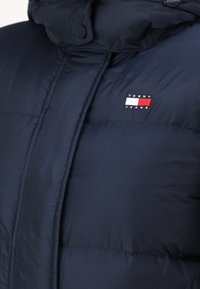 Jūrų laivyno pūkinė striukė su didelėmis kvadratų siuvinėtomis detalėmis. Krūtinėje yra mažas siuvinėtas Tommy Jeans logotipas raudonai, baltai ir mėlynai.