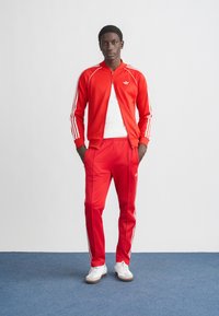 Fato de treino vermelho feito de tecido elegante, com riscas brancas nas mangas e nas calças, com um casaco com fecho de correr e um top branco justo por baixo.