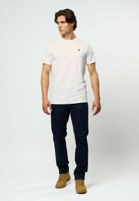 Balta trumpa rankovėmis marškinėliai su mažu tamsiu logotipu, derinami su tamsiai mėlynomis džinsomis ir šviesiai rudos spalvos batus su nėriniais, stovint neutralioje aplinkoje.