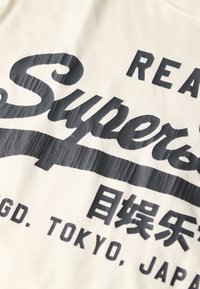Camiseta de algodón color crema con un atrevido texto impreso en negro, incluyendo "Super" en una escritura fluida y texto adicional en un estilo de bloque.