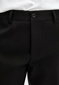 Gros plan d'une taille de pantalon noir ajusté avec bouton, passants de ceinture et braguette avant, porté avec une chemise blanche rentrée.