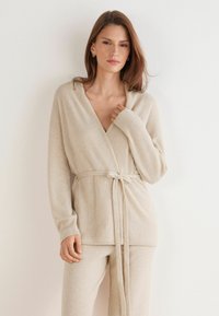 Cárdigan de punto beige con escote en V, mangas largas y cinturón atado. Presenta una textura suave y detalles acanalados en los bordes. Combinado con pantalones a juego.