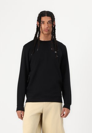 Sort sweatshirt med lange ærmer og rund hals. Har et diskret logo på brystet. Matcher med lyse beige, løstsiddende bukser.