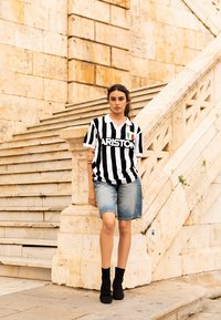 Camiseta deportiva a rayas en blanco y negro con el texto "ARISTON", combinada con pantalones cortos de mezclilla azul claro y botas negras hasta el tobillo, de pie en unos escalones de piedra.