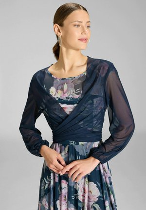 Vrouw met haar in een knot draagt grote creoolse oorbellen, een doorschijnende marineblauwe wikkeltop over een bloemenjurk, poseren met samengevouwen handen ter hoogte van de taille.