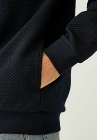 Navyblauwe sweatshirt met lange mouwen van textuurstof, diamantpatroon, geribde manchetten en zijzak, gedragen over lichte denim.