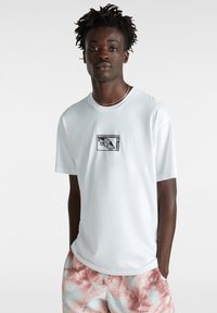 Camiseta blanca de manga corta hecha de algodón, con un parche de logo a cuadros negros en el pecho. Combinada con pantalones cortos de tie-dye en tonos pastel.