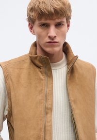 Rjava semiš brezrokavnik z visokim ovratnikom, sprednjo zadrgo in stranskimi žepi, nošen preko svetlega rebrastega puloverja. Gladek zaključek, minimalističen dizajn.