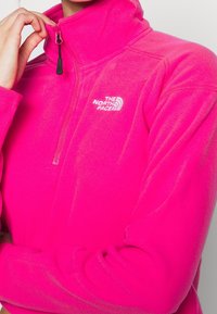 Różowy polarowy półgolf z wysokim kołnierzem, przednim suwakiem w połowie długości i odblaskowym białym logo. Miękka tekstura i sportowy design.