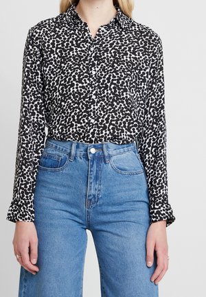 Mujer con una camisa de botones con un patrón abstracto en blanco y negro metida en unos vaqueros azul de tiro alto, de pie frente a un fondo liso.